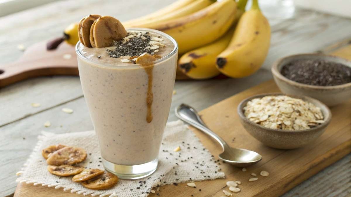 A high-protein milkshake in a glass, topped with chia seeds and almond butter, surrounded by oats, bananas, and protein powder on a wooden kitchen counter